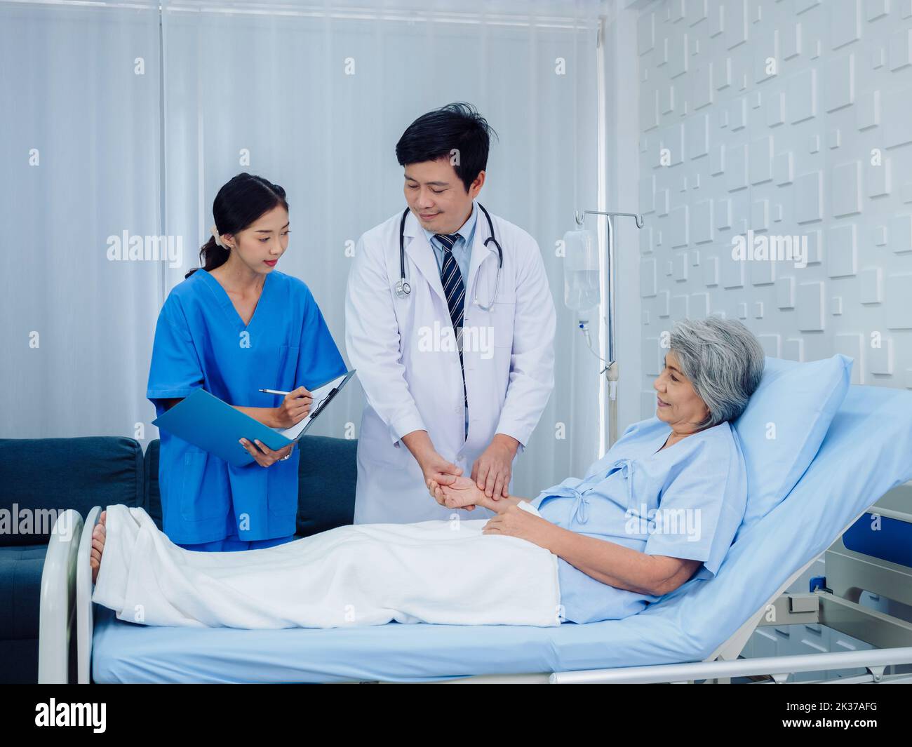 Asian male doctor, professional in white lab coat holding elderly patient's hand for check to examine heartbeat pulse and nursing assistant taking not Stock Photo