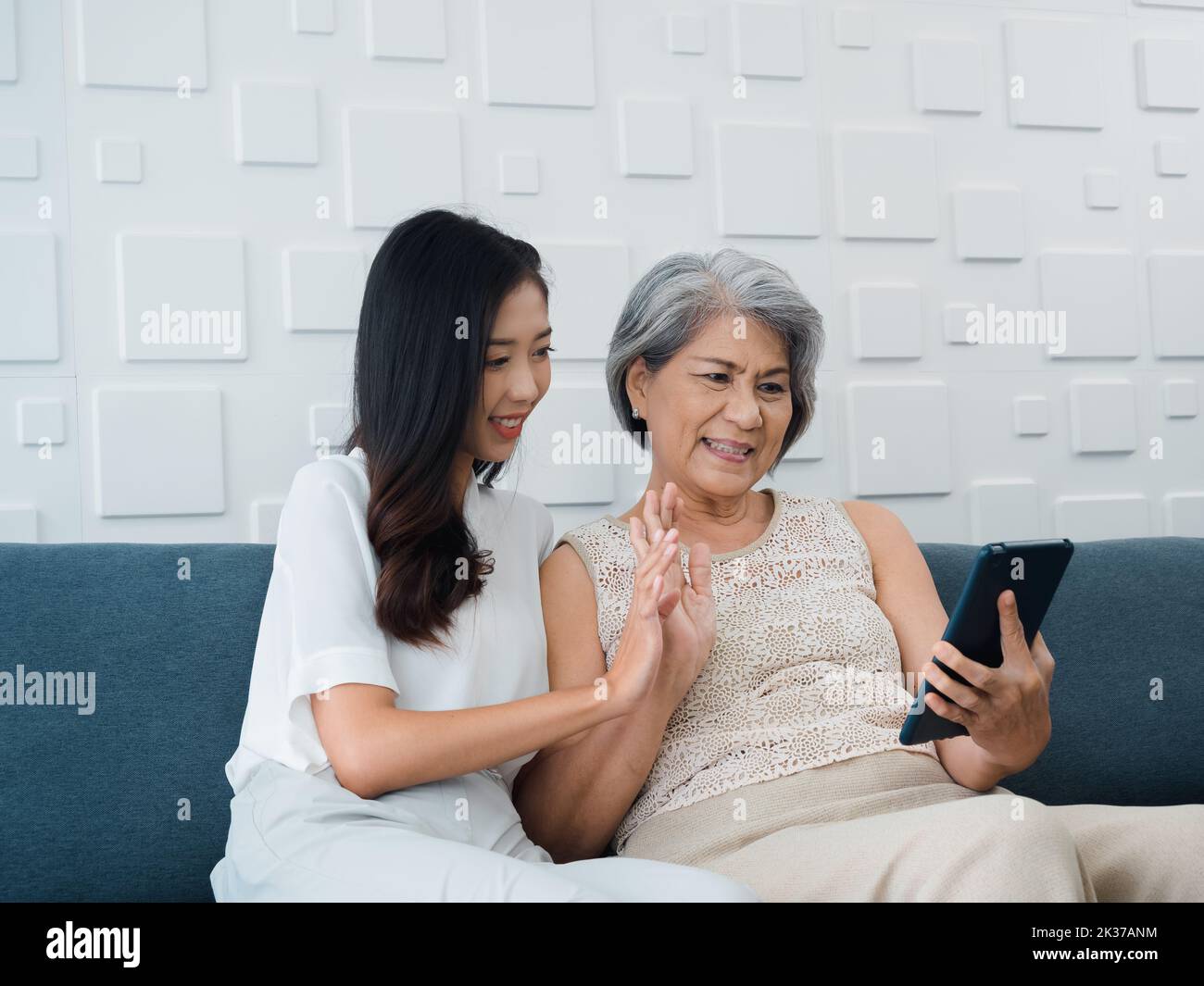 Happy lovely Asian senior woman, mother white hair and beautiful young female, daughter waving greeting at digital tablet screen while sit on couch in Stock Photo