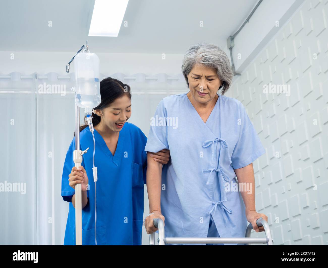 Elderly Asian woman patient trying to walk on walking frame held and carefully supported in arms by caregiver, young polite female assistant nurse in Stock Photo