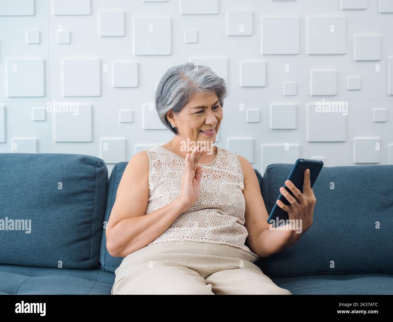 Happy Asian senior casual woman sitting on couch, waving greeting at digital tablet screen in hand in white room. Elderly female meet with family by v Stock Photo