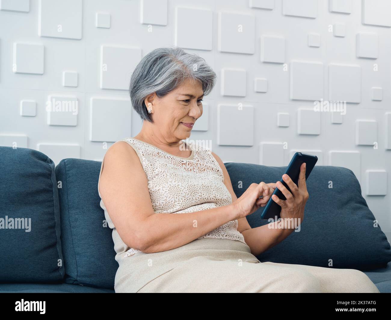 Happy Asian senior woman sit on couch, holding and looking at digital tablet screen in hand in living room. Elderly female using smart device, compute Stock Photo
