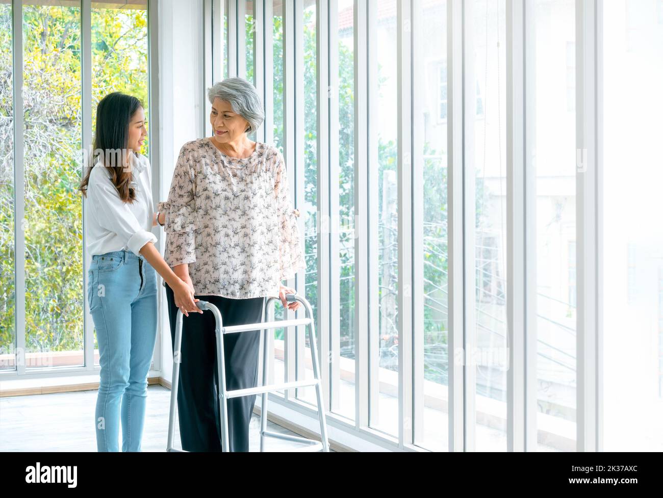 Caregiver, smiling young Asian female, daughter or grandchild supporting help elderly woman, mother or grandparents try to walk with walker frame at h Stock Photo