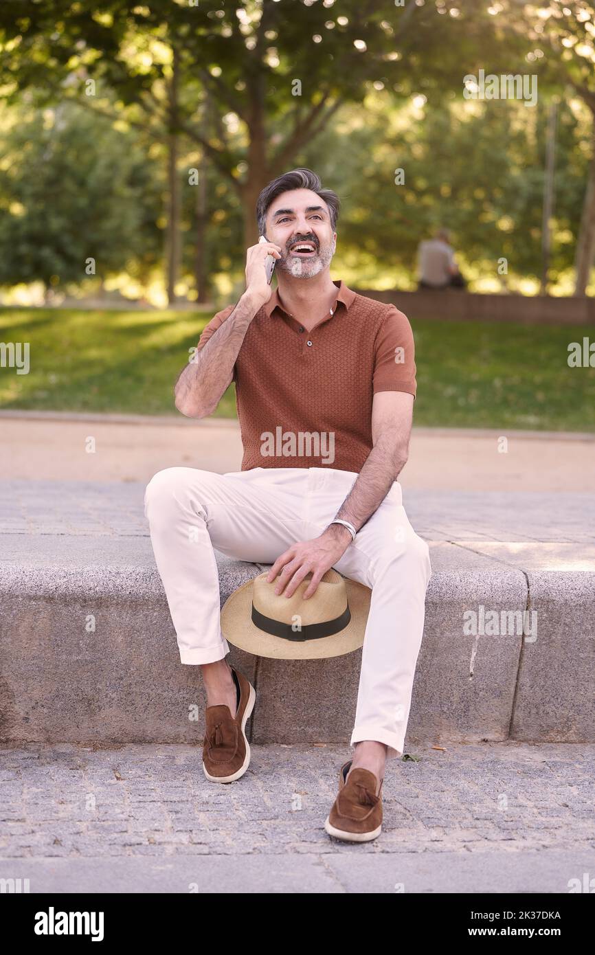 Relaxed man with a hat in hand smiling while talking on the phone sitting outdoors in a park. Technology concept. Stock Photo