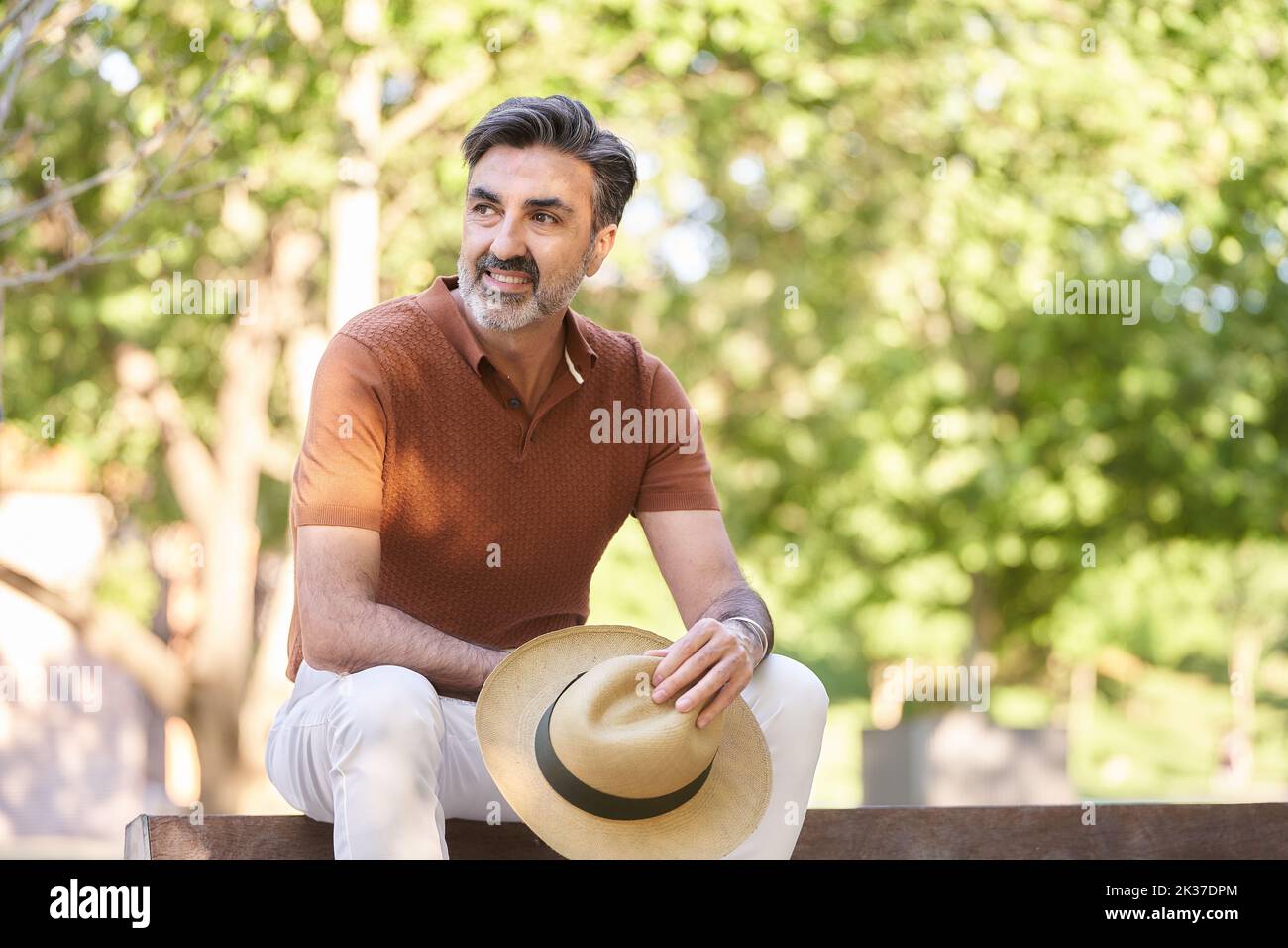 Relaxed middle-aged man holding a hat and looking away while sitting on a bench in a park. Stock Photo