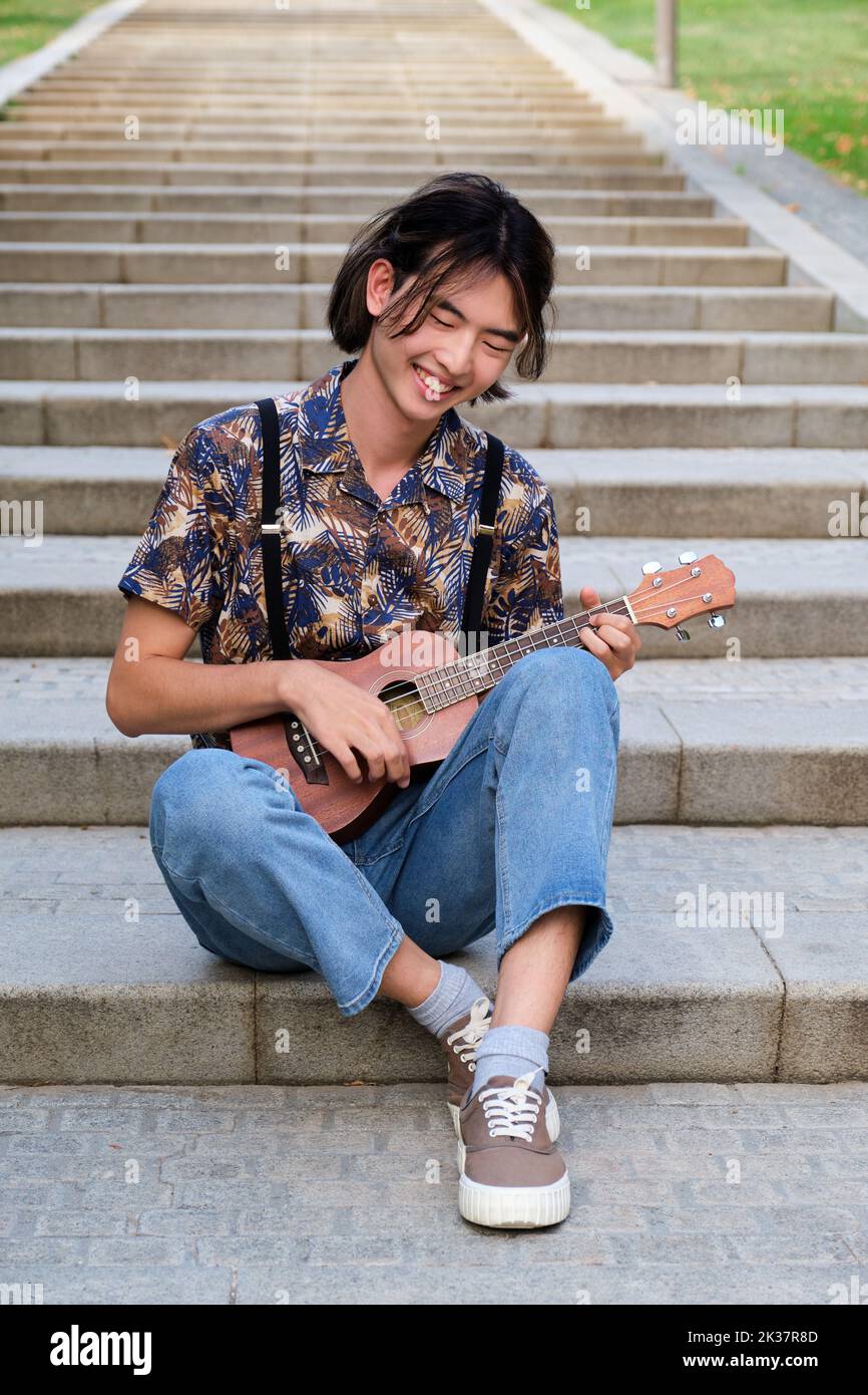 Taiwanese guy smiling and playing acoustic Ukulele guitar sitting on stairs. Stock Photo