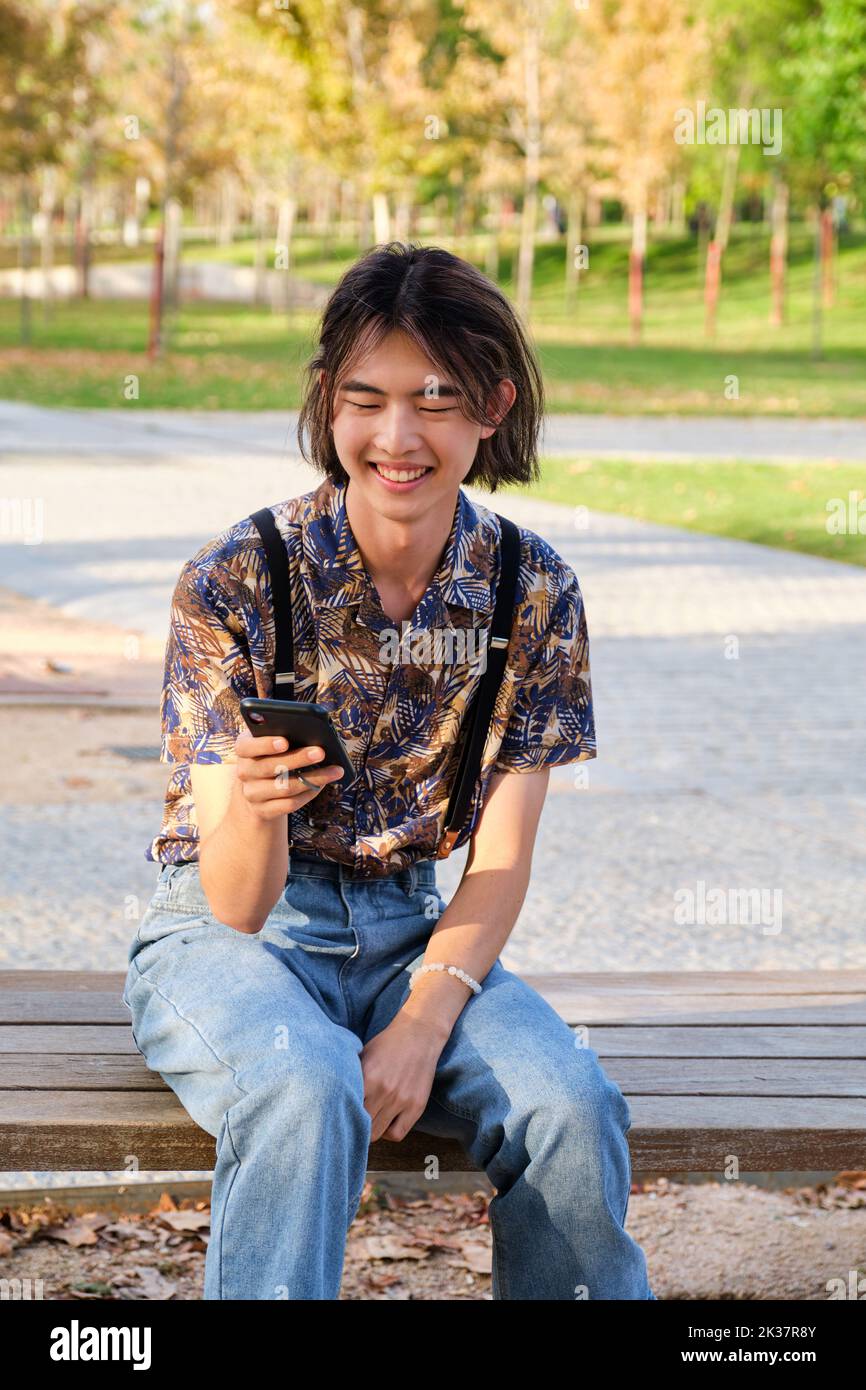 Asian guy laughing and using the smartphone sitting a bench. Stock Photo