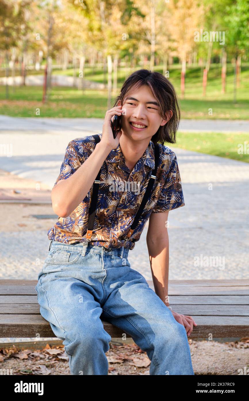 Taiwanese guy smiling and talking on the smartphone sitting a bench. Stock Photo