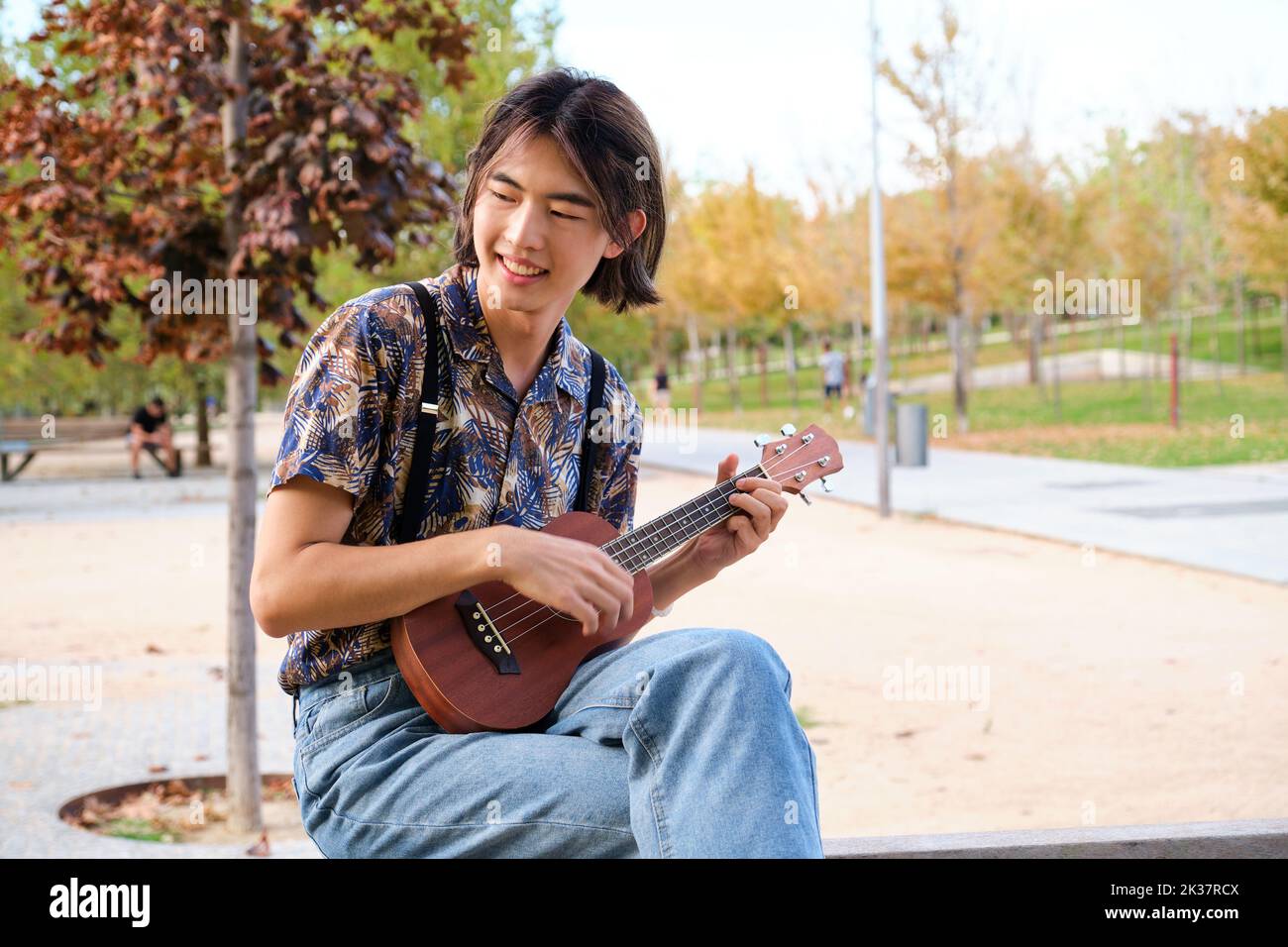 Taiwanese guy smiling and playing acoustic Ukulele guitar sitting on a bench. Stock Photo