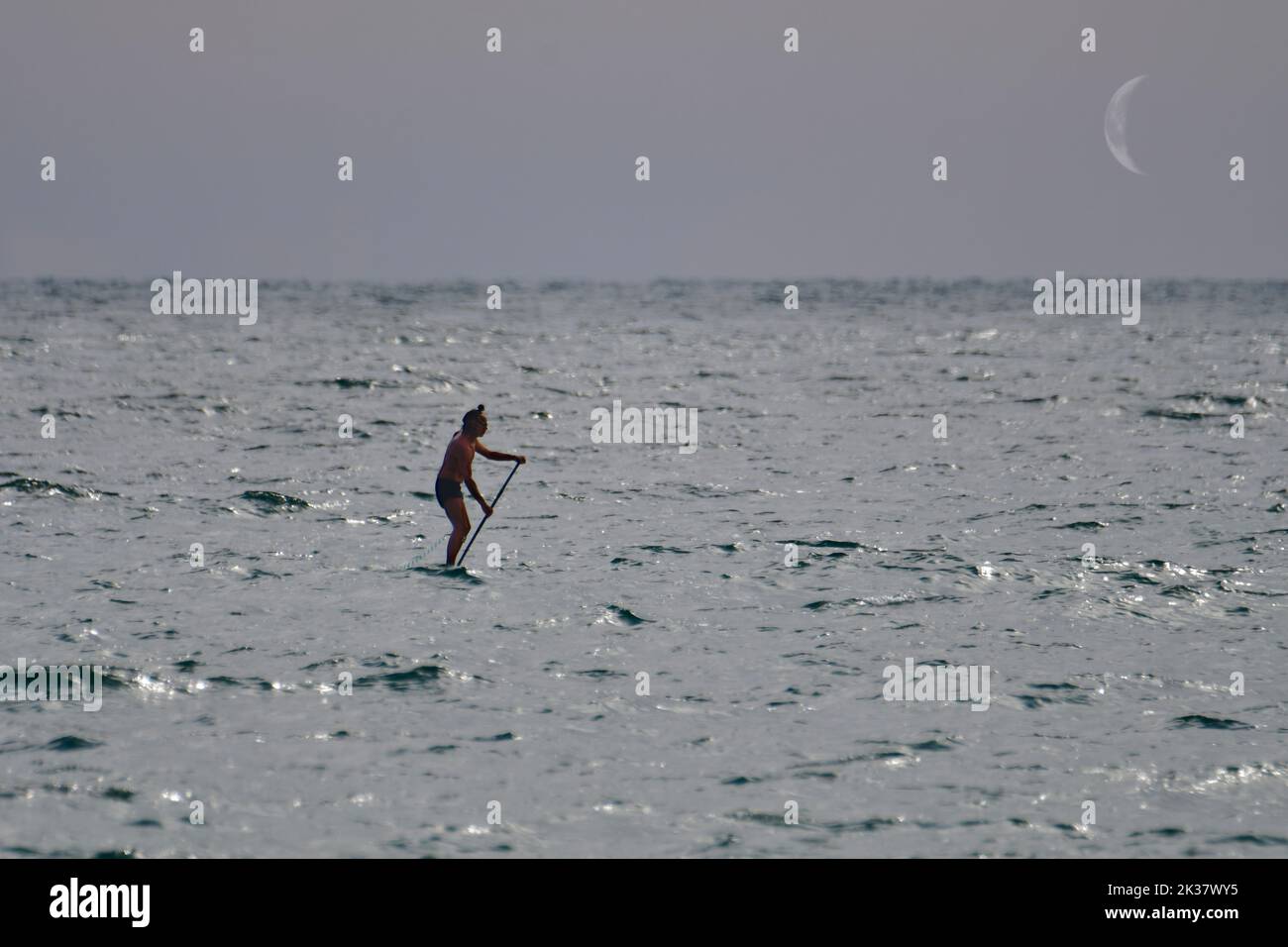A man practices paddle surfing on a hot day with the Moon as a witness. Stock Photo
