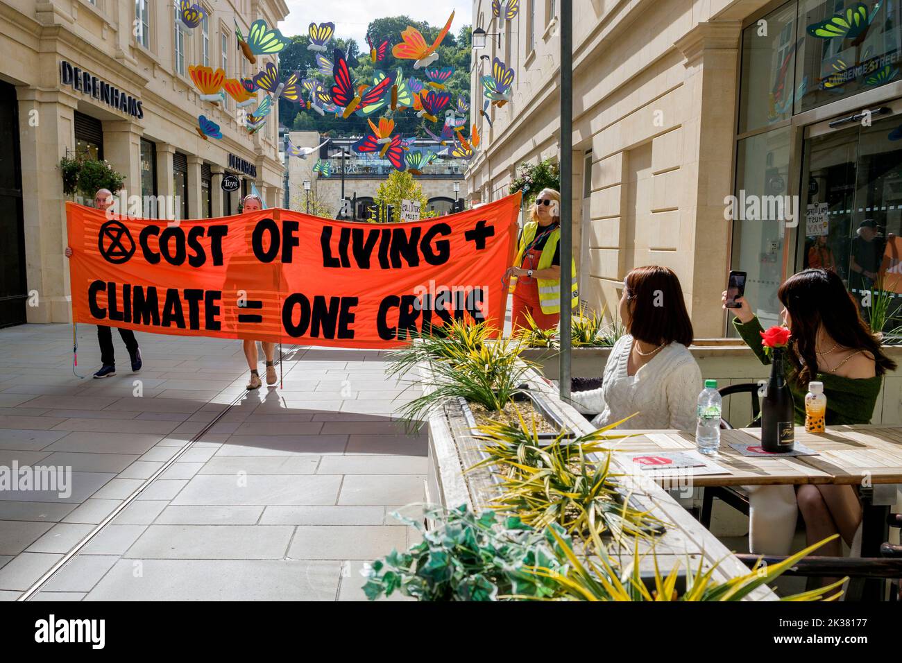 Bath, UK. 25th Sep, 2022. With PM Liz Truss signalling an acceleration of oil and gas extraction in the UK shoppers enjoying a drink look on as climate change protesters take part in a protest march through the centre of Bath. The protest organised by Extinction Rebellion was held in order to highlight how the cost of living crisis and the climate crisis are interlinked. Credit: Lynchpics/Alamy Live News Stock Photo