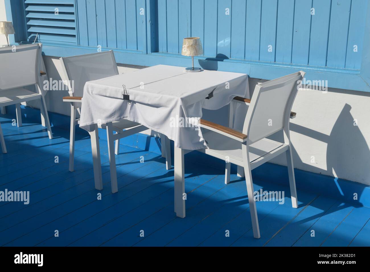 White table and chairs with blue wooden floor and wall as backdrop. Outdoors in sunshine with shadows. Restaurant, no people. Stock Photo
