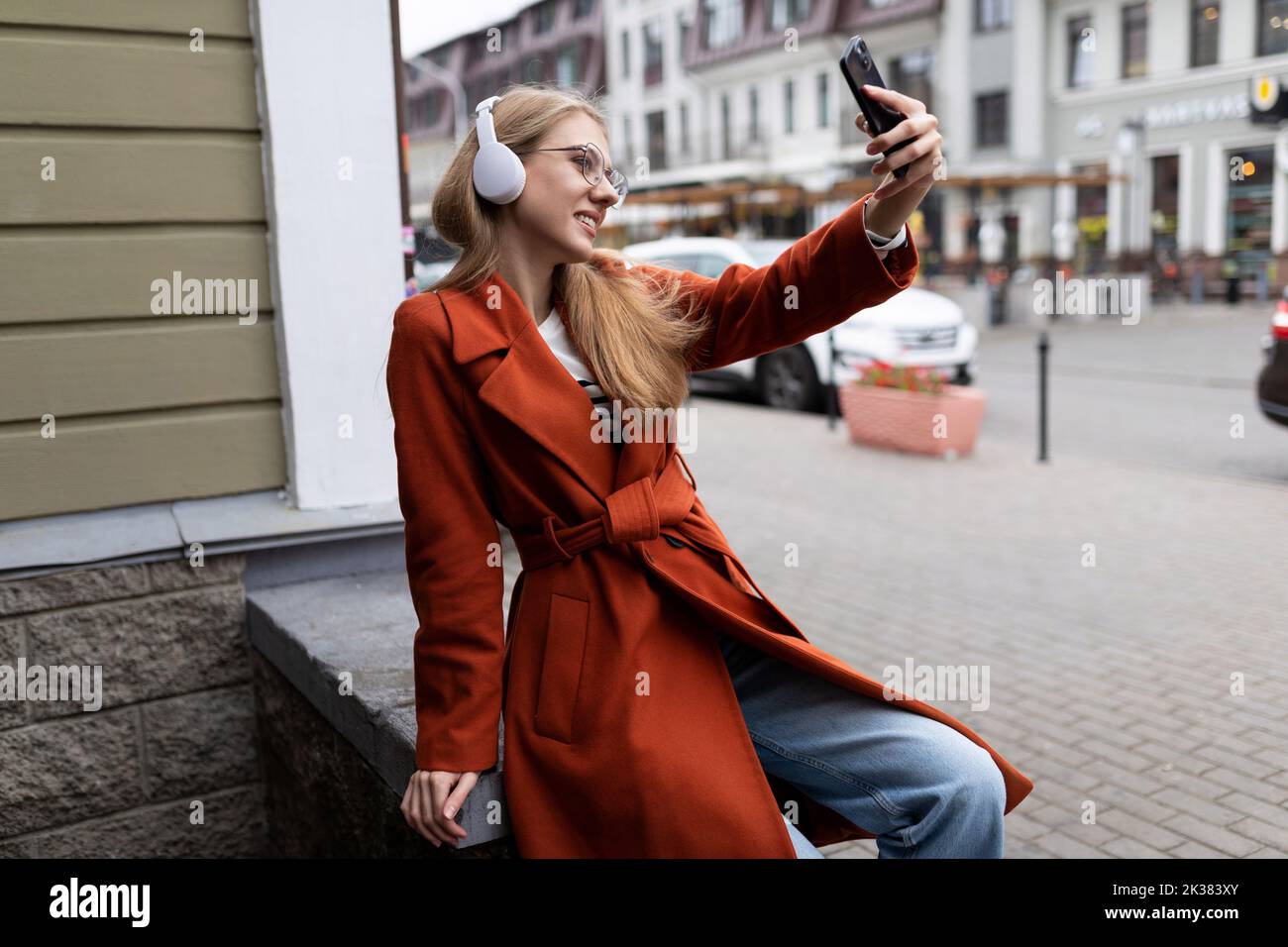 a young female student in a red coat and headphones takes a selfie ...