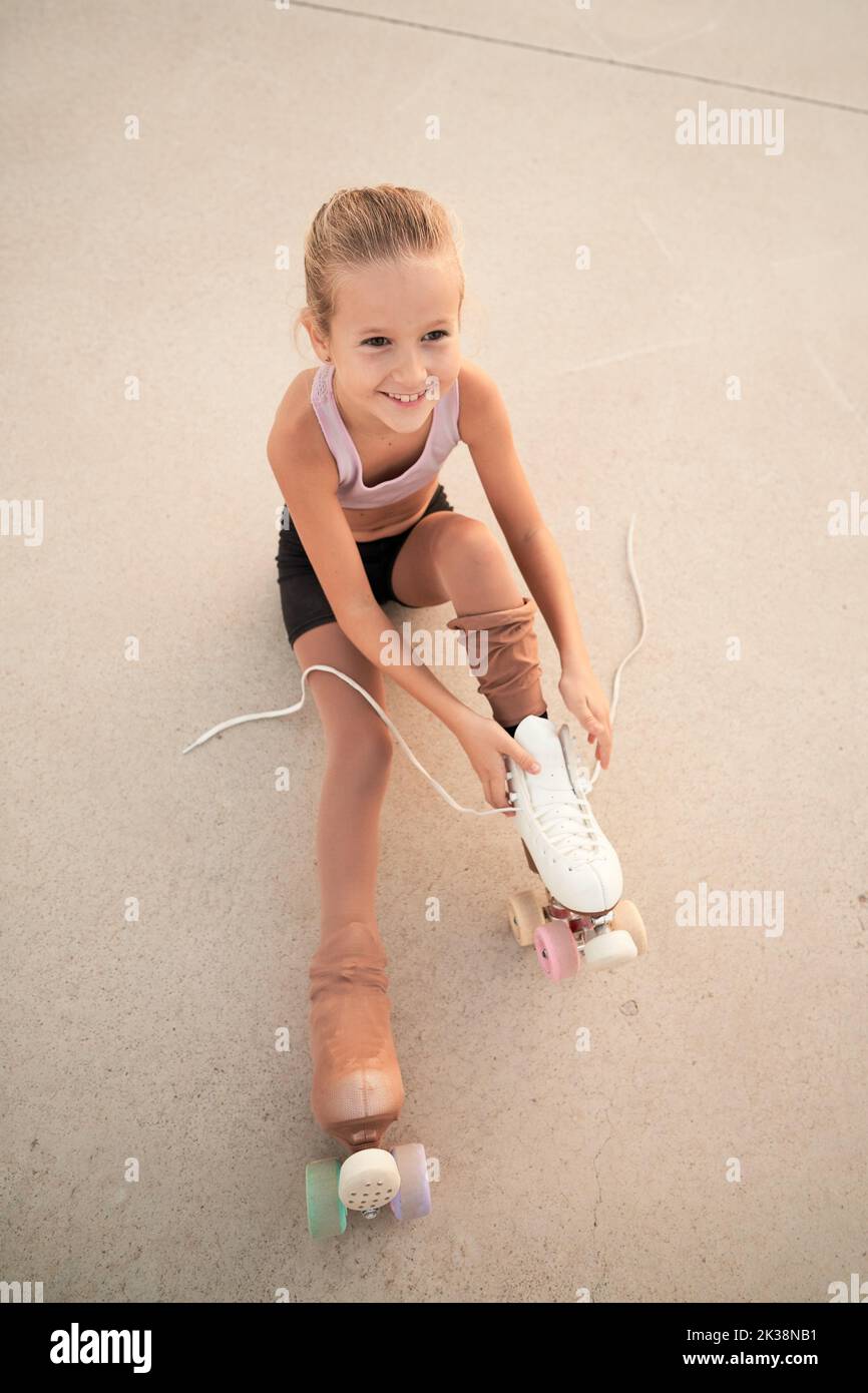 Child lacing up roller skates while sitting on concrete ground Stock