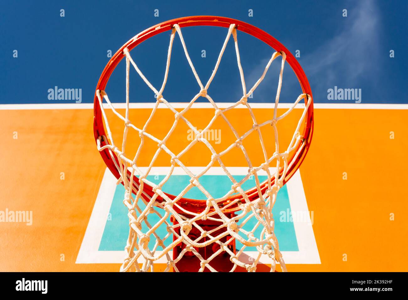 Low angle view of colourful orange basketball backboard with red hoop and a net against blue sky. Stock Photo