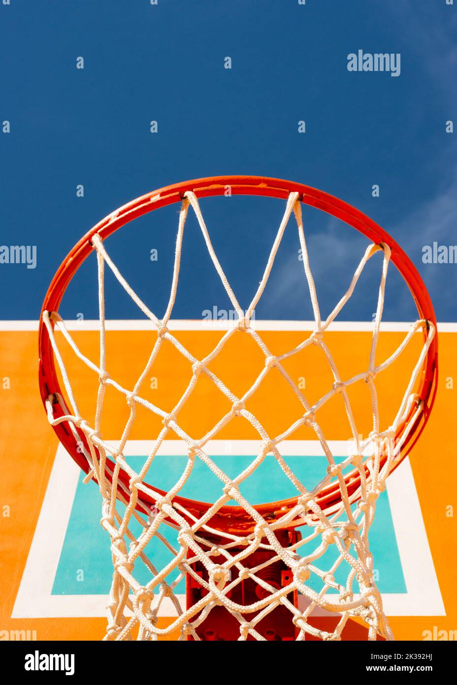Colorful orange basketball backboard with red hoop and a net against blue sky low angle view. Stock Photo