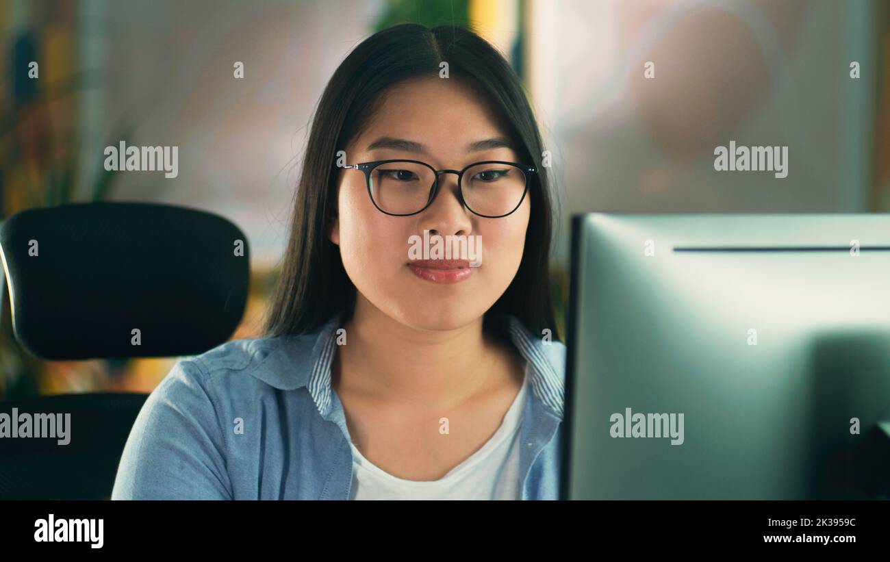 Asian woman in glasses looking at screen of computer while working on 3D modeling project or design remotely from home or surfing the internet in spare time. Freelance Stock Photo