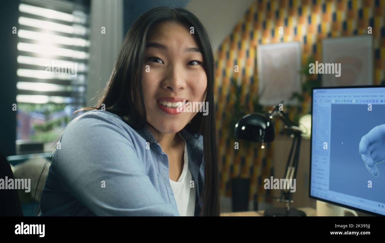 Surprised asian woman rejoicing and looking at camera while sitting at the table at home office and working remotely on 3D modeling project on computer Stock Photo