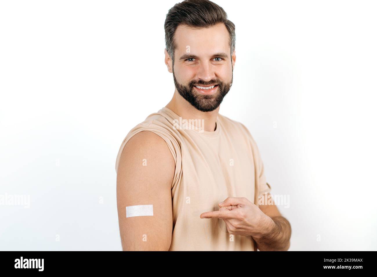 Vaccination, disease prevention. Positive joyful caucasian man, with a plaster on his shoulder, received a vaccine, points a finger at it, stands on a white isolated background, looks at camera, smile Stock Photo