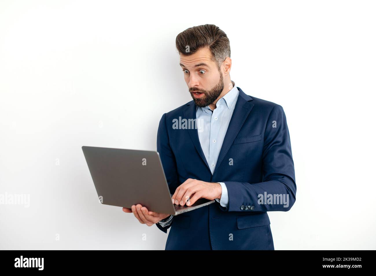 Shocked surprised caucasian bearded business man, in a suit, programmer, IT specialist, holding opening laptop in hands, looks in amazement at the screen, standing on isolated white background Stock Photo