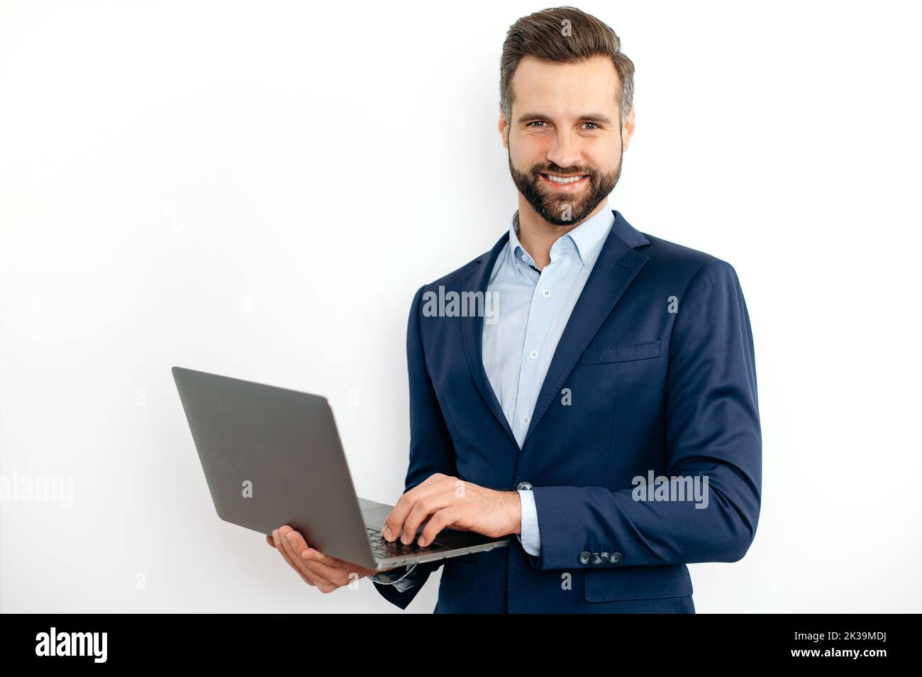 Friendly attractive caucasian bearded business man, in a suit, programmer, IT specialist, holding opening laptop in hands, standing on isolated white background, looks at camera, smiling Stock Photo
