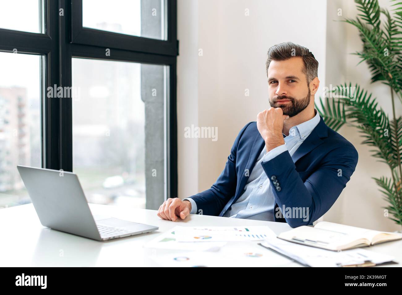 Photo of positive handsome elegant caucasian man, entrepreneur, company ceo, sitting at workplace in the office, dressed in formal suit, looking at camera. Attractive successful bearded businessman Stock Photo