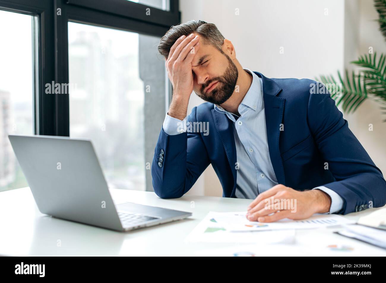 Stressed worried upset tired caucasian businessman, corporate director, financial manager, sits in the office at workplace, nervous of deadline a project, closed his eyes, holding hand on head Stock Photo