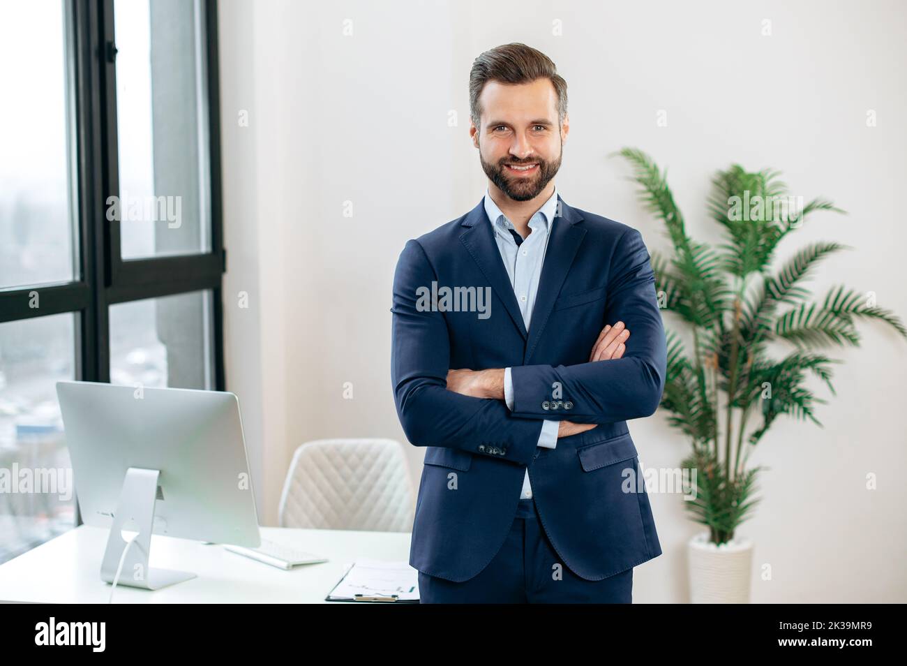 Portrait of a handsome confident elegant successful caucasian bearded man, in a suit, top manager, company ceo, financial director, stand in the office, arms crossed, looking at the camera, smiling Stock Photo