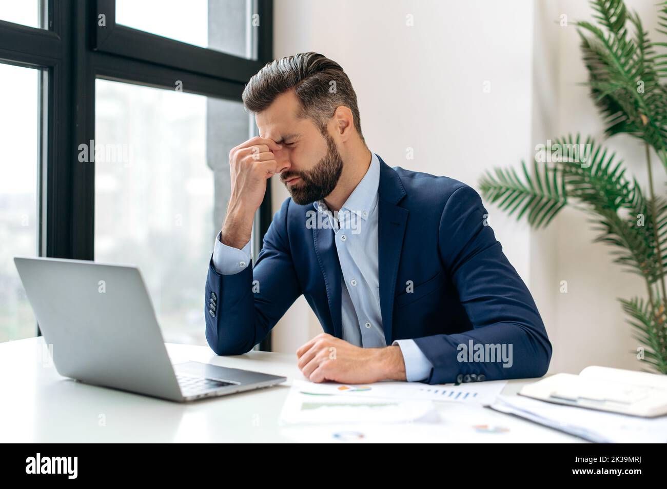 Unhappy tired caucasian businessman, corporate manager, sits at the workplace, tired from online work, stressed, nervous, massages the bridge of his nose, closed his eyes, experiencing a headache Stock Photo