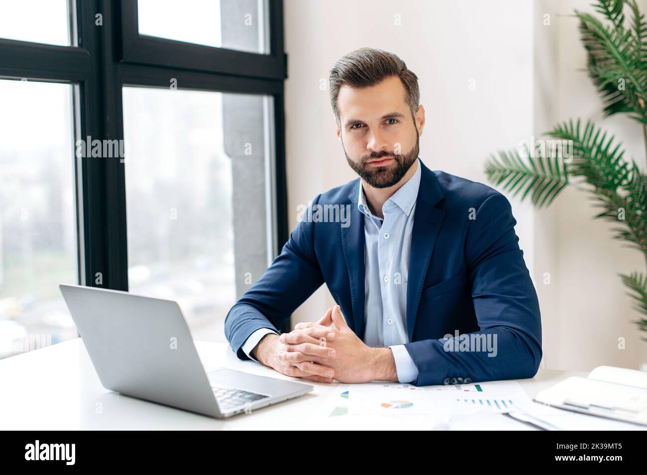 Portrait of a successful confident attractive influential caucasian bearded businessman in a formal elegant suit, ceo company, sits at a work desk in the office, looking at the camera Stock Photo