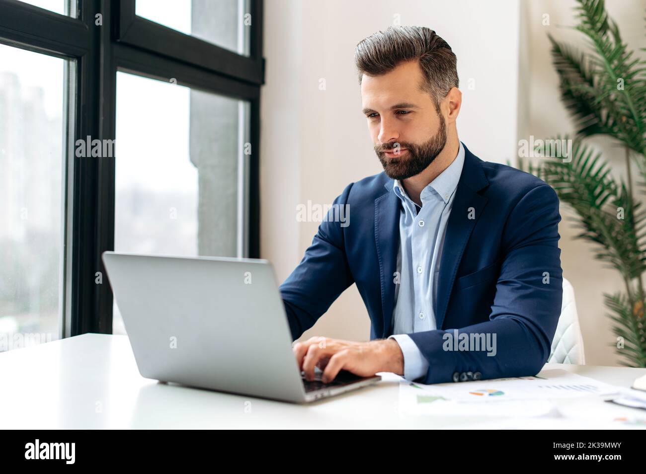 Successful confident busy caucasian bearded businessman in a formal elegant suit, sits at workplace, working on a project uses laptop, writing business strategy, conducts working correspondence online Stock Photo