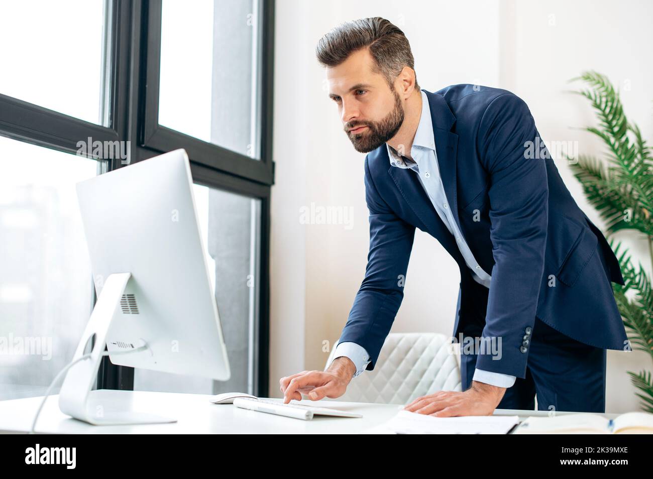 Busy concentrated confident caucasian bearded man, company ceo, top manager, stand near work desk in the office, working in computer, conducts working correspondence online, views information Stock Photo