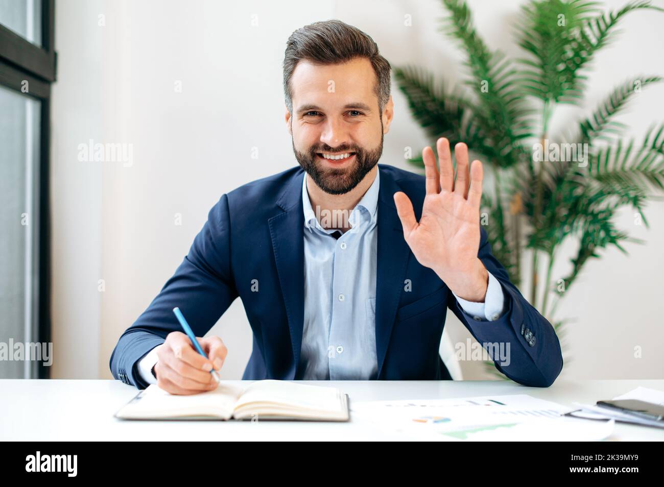 Positive caucasian professional business man, mentor, waving hand and looking at webcam during video conference, listens lecture, leading webinar, record online training, smiling friendly Stock Photo