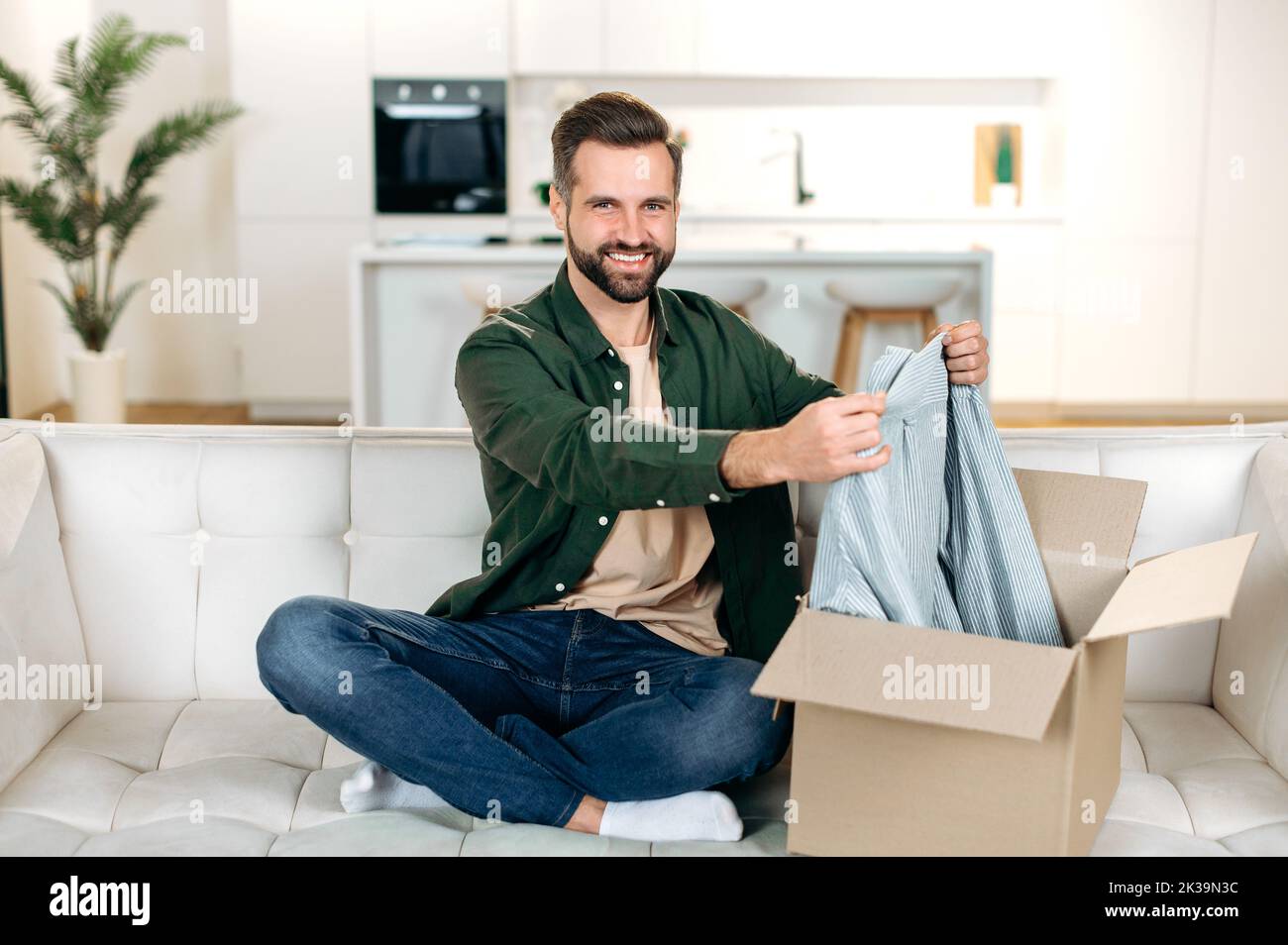 Handsome positive caucasian man opening big carton box while sits on sofa, unpacking internet store order at home, feel satisfied with fast delivery service and the goods which received, smile happily Stock Photo