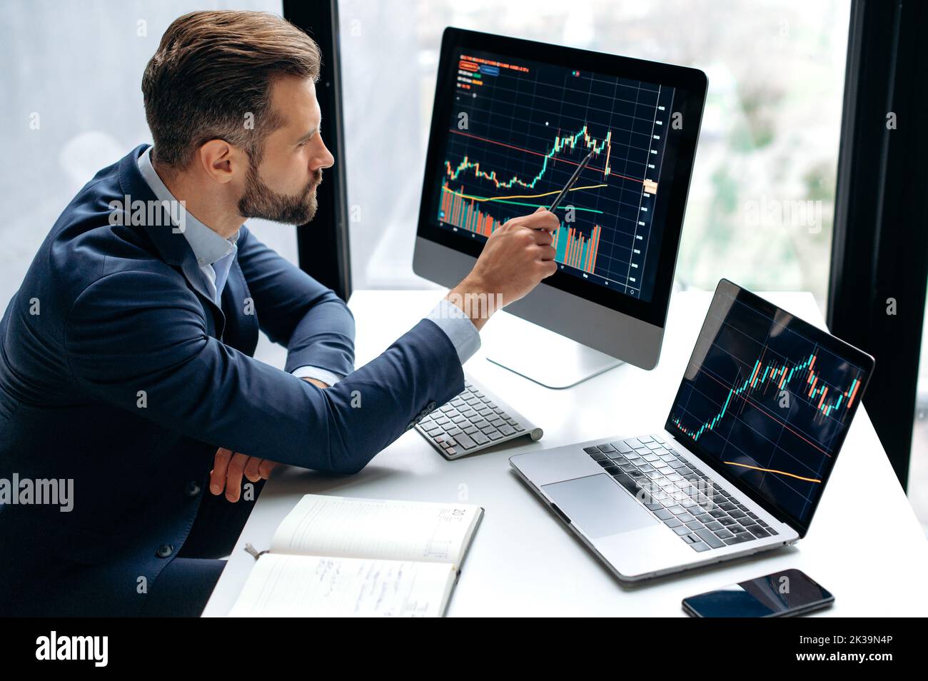 Focused smart successful caucasian male stock investor, broker, sits at his work desk, uses computer and laptop, concentrated analyzes risks and prospects, rise or fall of cryptocurrency coin Stock Photo