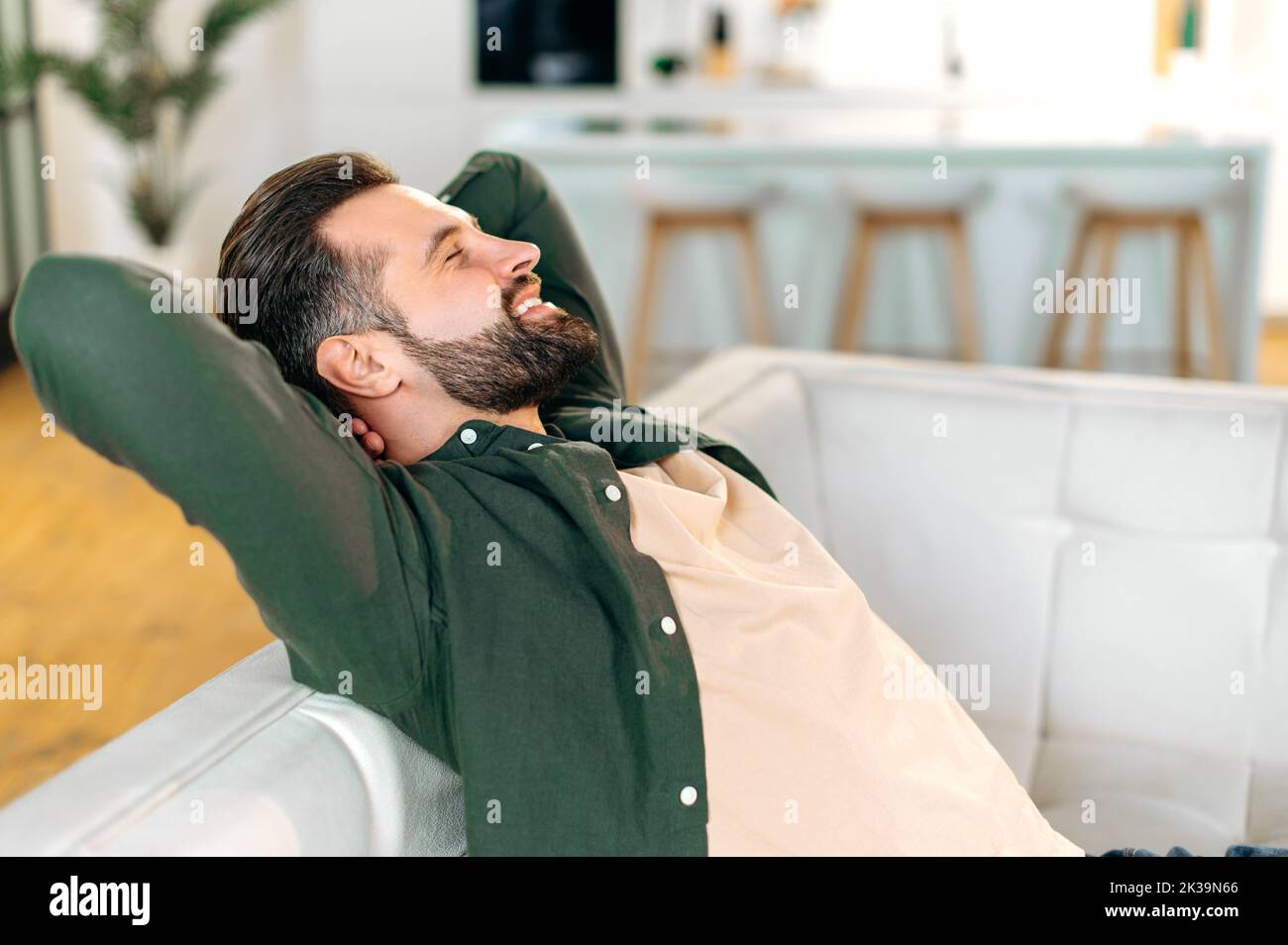 Satisfied calm happy caucasian bearded man, in a casual shirt, sits on a sofa in the living room with his hands behind his head, closing his eyes, relaxes after a working day, rests, smiles Stock Photo