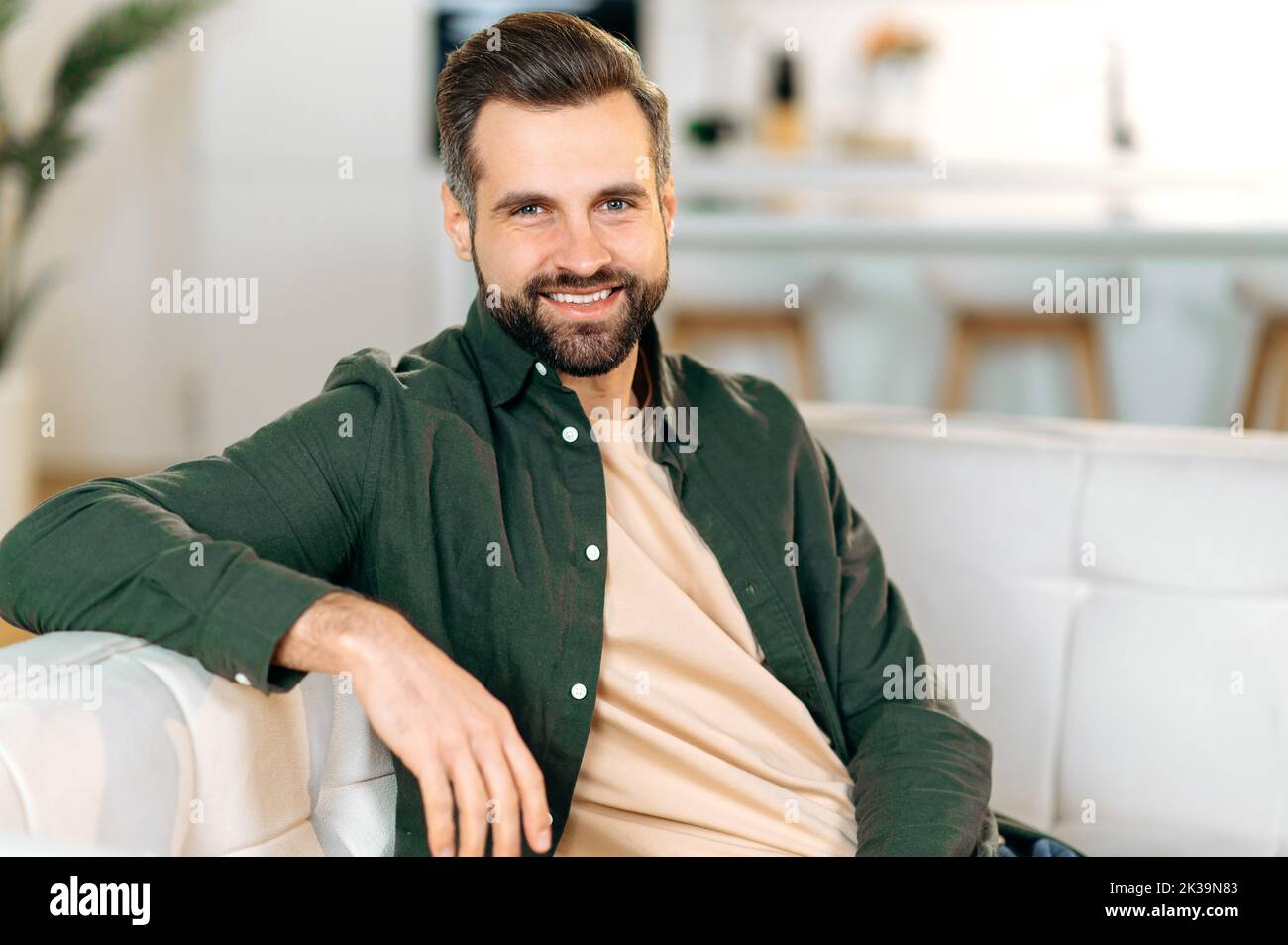 Portrait of a handsome attractive modern bearded caucasian man, in casual stylish clothes, sits on a sofa in the living room, looks at the camera, smiles friendly Stock Photo