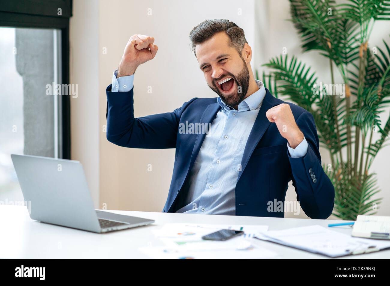 Victory concept. Successful cheerful lucky caucasian bearded businessman, ceo company, manager, sits at work desk in the office, gesturing with fists, celebrate success, win, big profit, job promotion Stock Photo