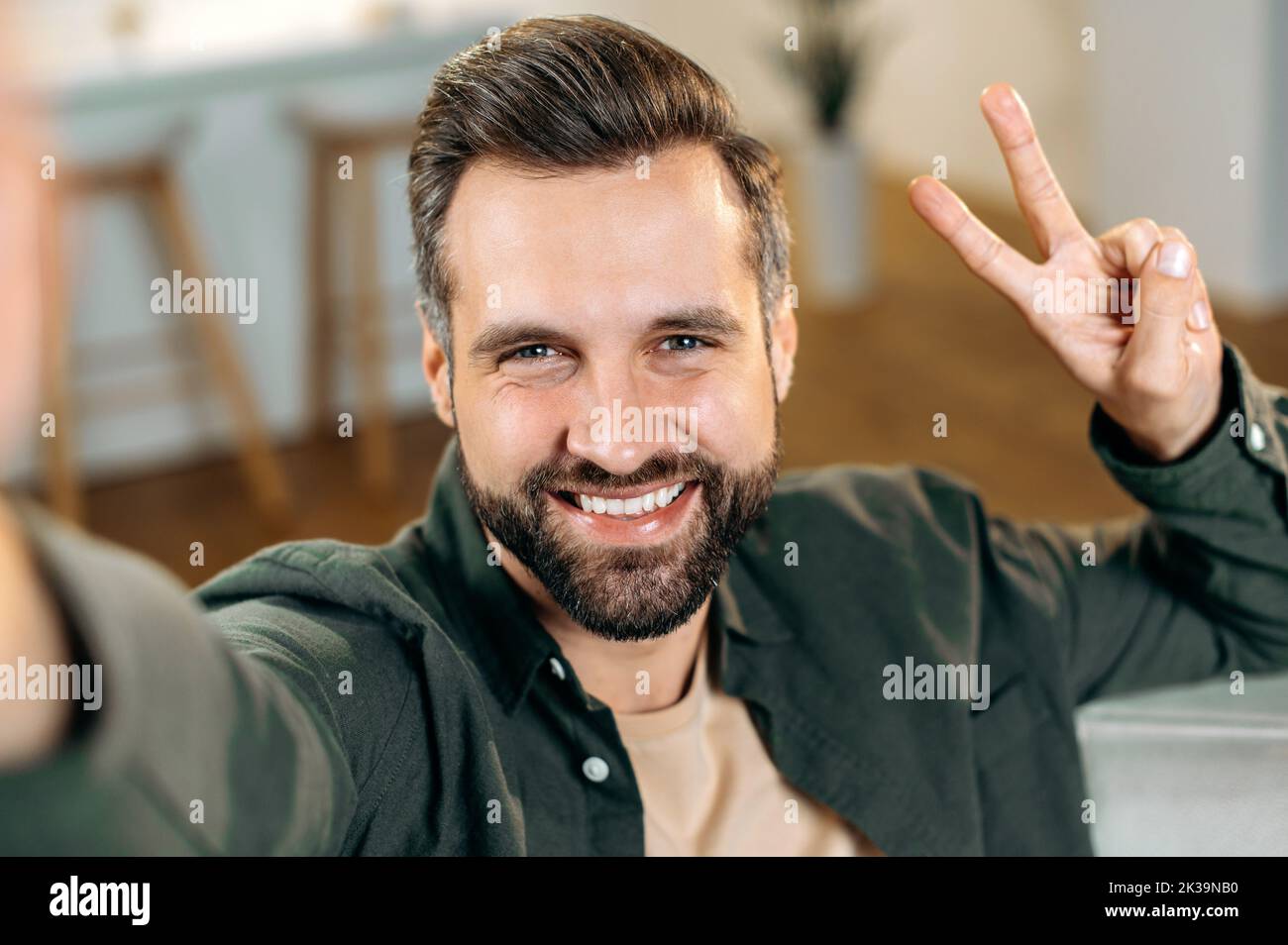 Joyful handsome positive caucasian bearded man, in a casual shirt, takes a selfie on his smartphone, shows a peace sign with fingers, looks into the front camera of the phone, smiles friendly Stock Photo