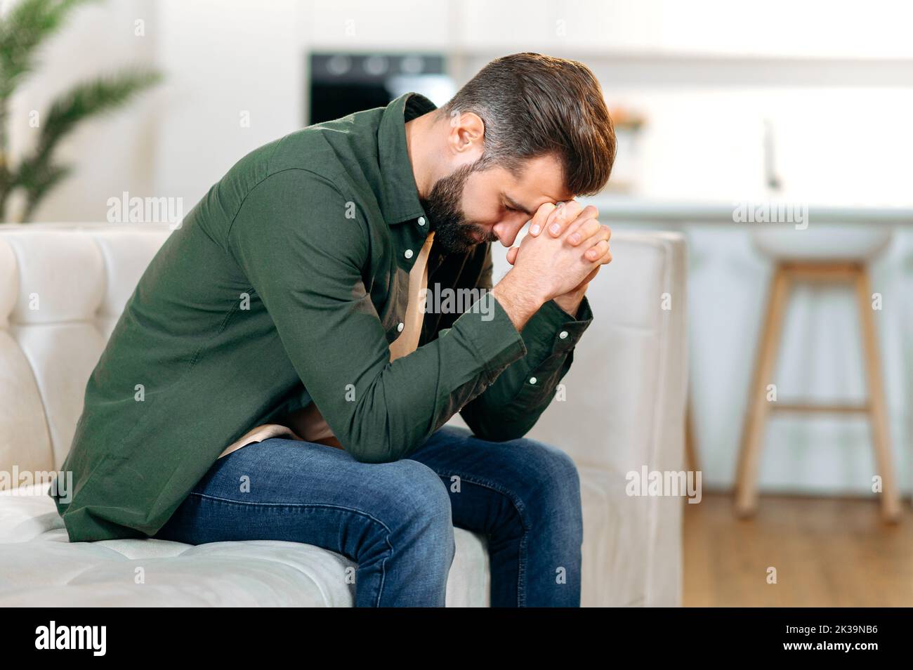 Upset frustrated caucasian man, sitting on the sofa in the living room with his head down and his eyes closed, experiencing personal problems, stress, loneliness, needs psychological support and rest Stock Photo