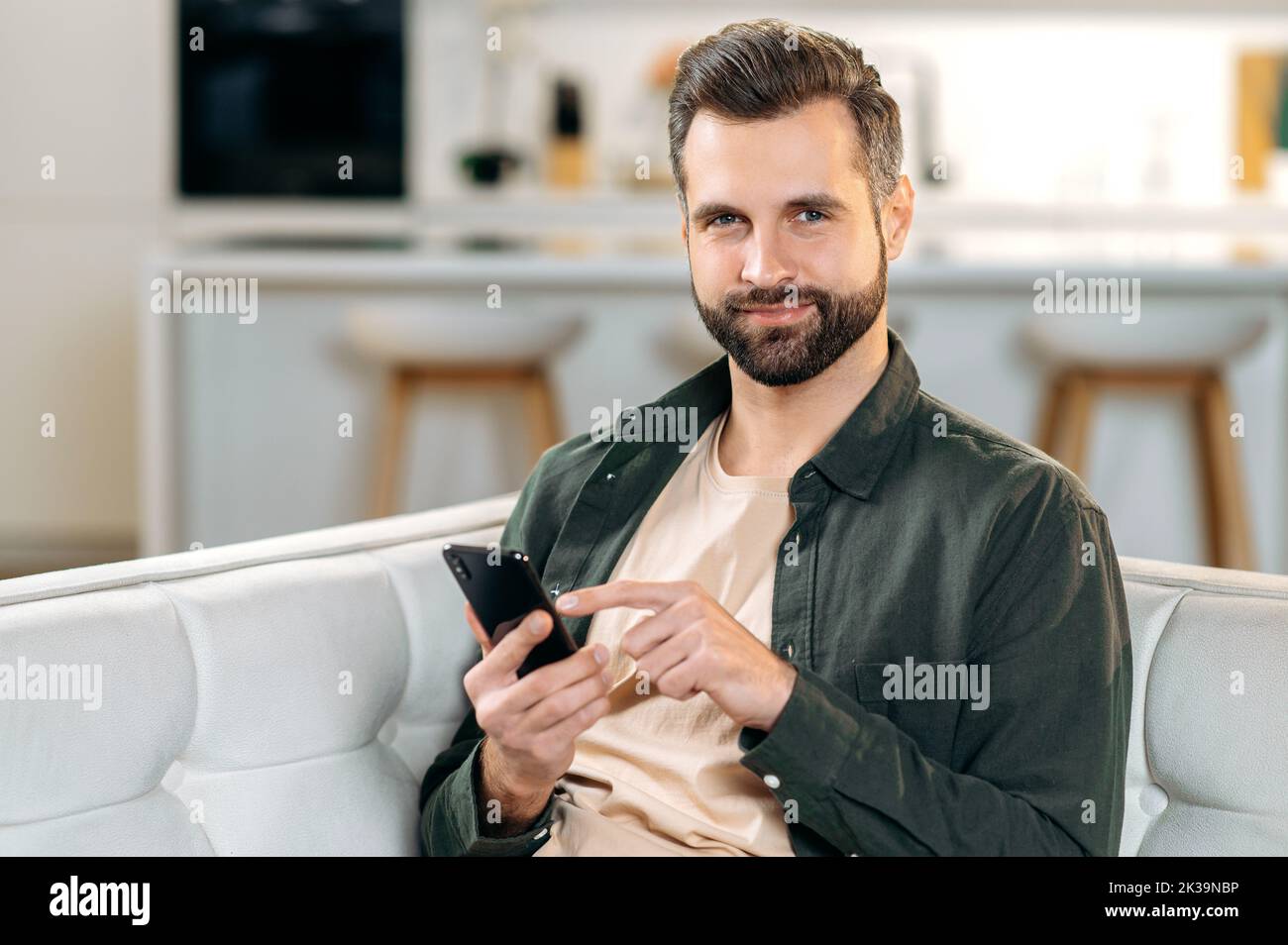 Positive attractive caucasian bearded stylish man, sitting at home on a sofa in a living room, using his smart phone, relaxing on couch, messaging online in social media, looks at camera, smiles Stock Photo