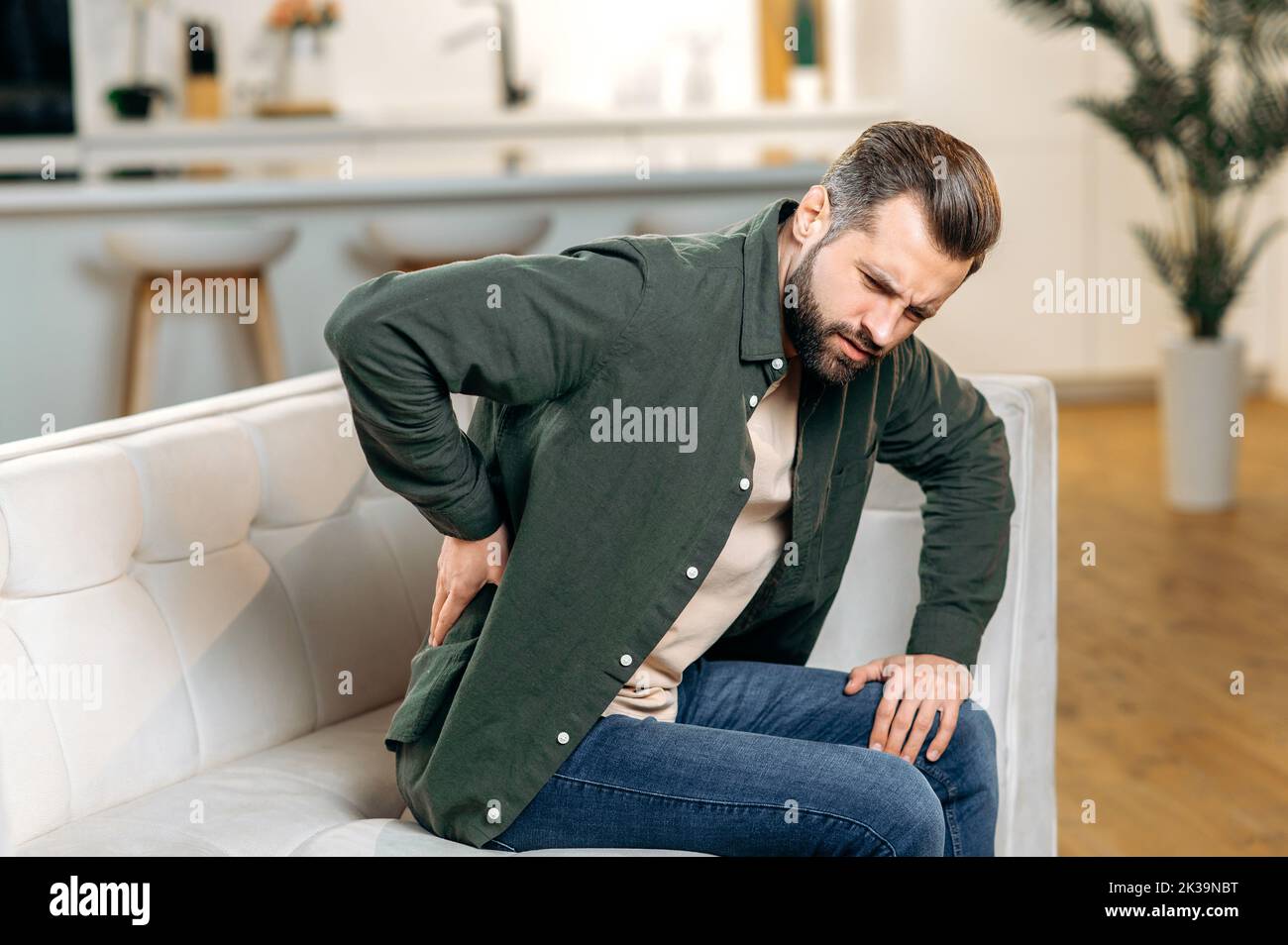 Unhappy caucasian man, dressed in casual wear, sitting on sofa in living room, experiencing acute pain in the back, lower back, has chronic joint problems. Problems with the back, spine, sciatica Stock Photo