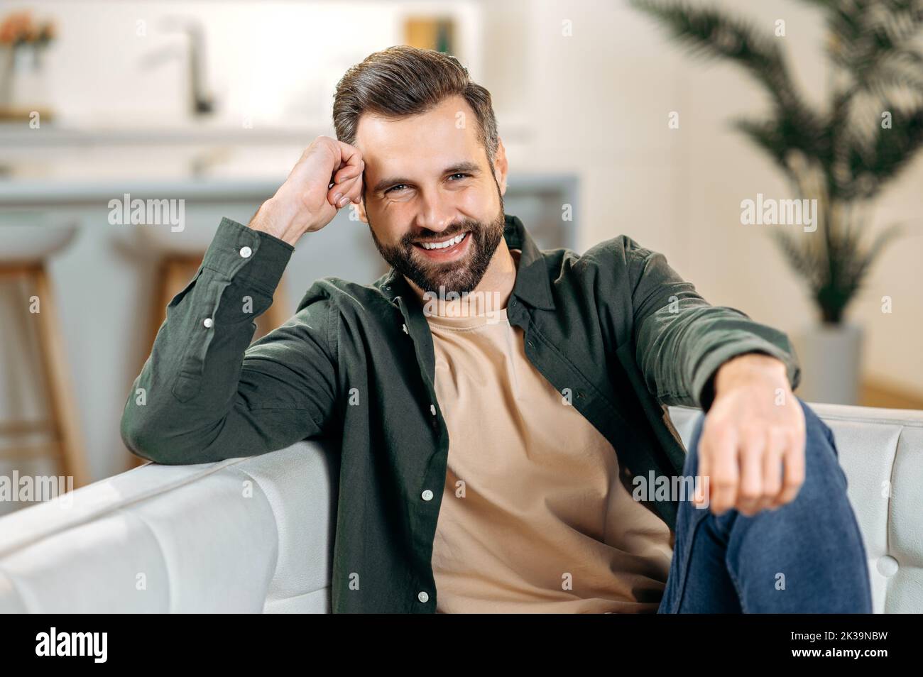 Male portrait. Headshot of a handsome attractive positive modern bearded caucasian man, in casual stylish clothes, sits on a sofa in the living room, looks at the camera, smiles friendly Stock Photo