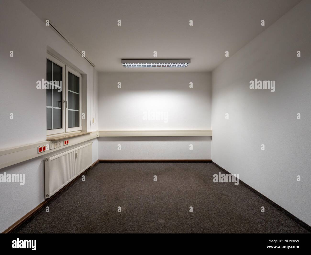 Real empty office room with dirt spots on the carpet. Old interior design in a commercial building. White walls with cable ducts. Fluorescent light. Stock Photo