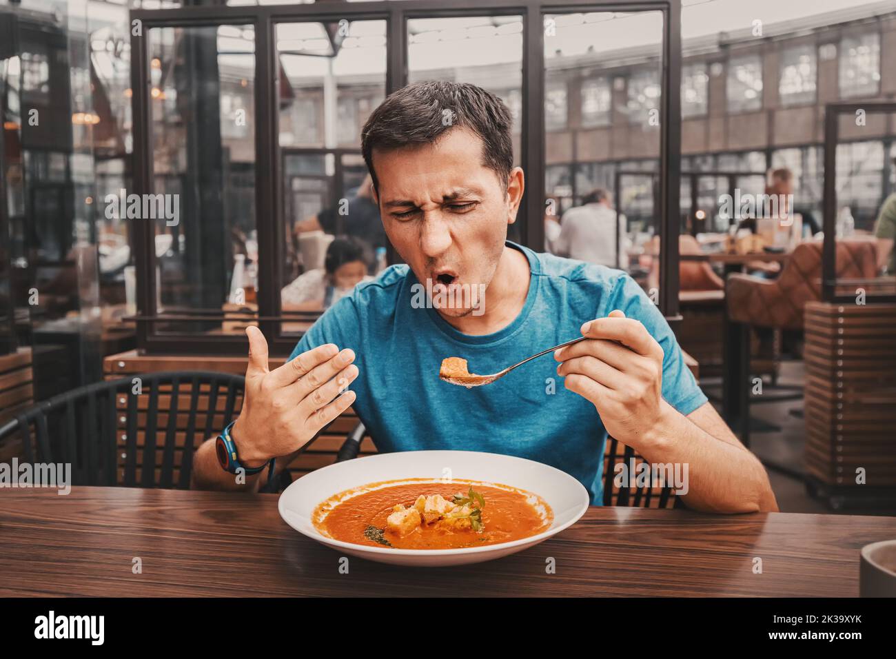 A man tries a spicy and hot red soup in a restaurant and reacts funny emotionally. Seasonings in the national cuisine and an unhealthy diet with overa Stock Photo