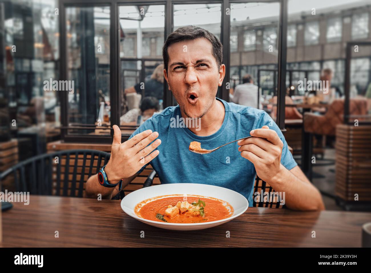 A man tries a spicy and hot red soup in a restaurant and reacts funny emotionally. Seasonings in the national cuisine and an unhealthy diet with overa Stock Photo