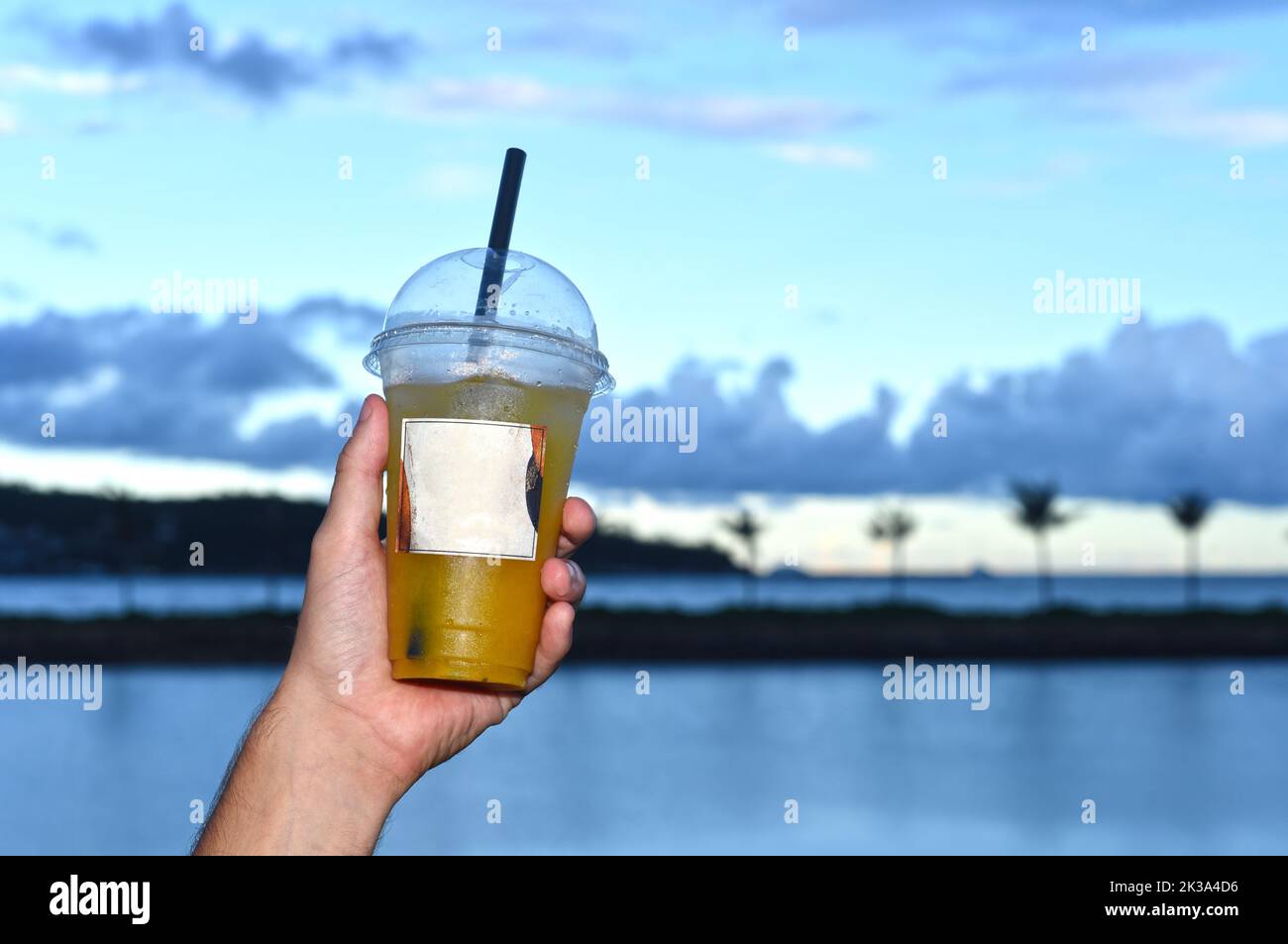Hand holding plastic cup with iced tea take away against sea and sunset Stock Photo
