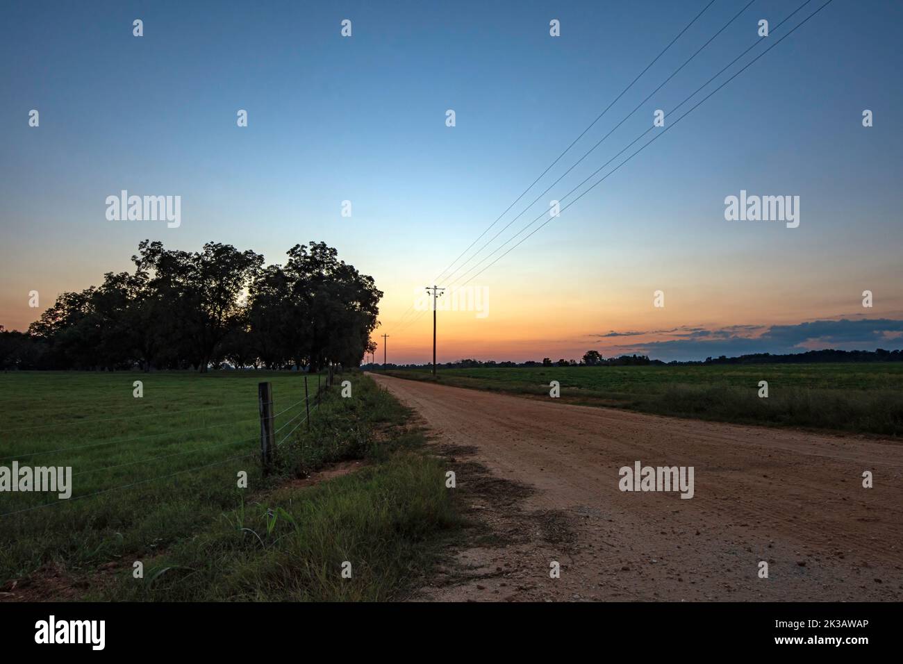 Background of a red dirt road leading into the sunset with a fenced pasture on the left and a peanut field on the right. Stock Photo