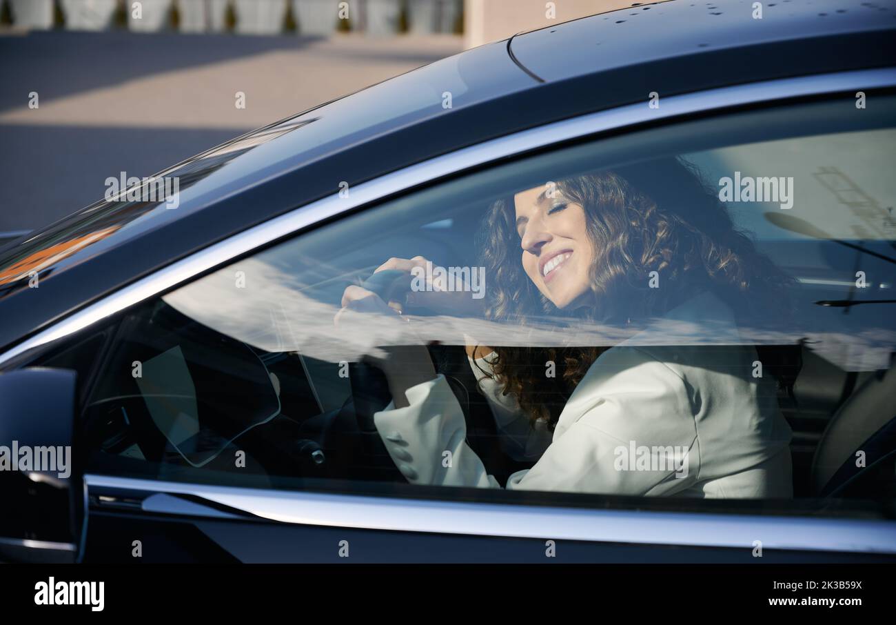 Beautiful businesswoman with closed eyes sitting in electric car and smiling while using vehicle autopilot system. Pretty woman holding steering wheel and sleeping while sitting on driver seat in car. Stock Photo