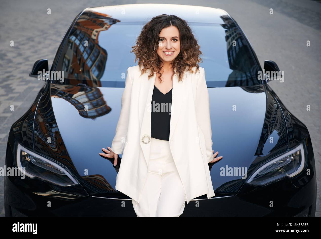 Portrait of young woman looking at camera and smiling while sitting on hood of electric car. Cheerful businesswoman in elegant white jacket posing near electric vehicle outdoors. Stock Photo