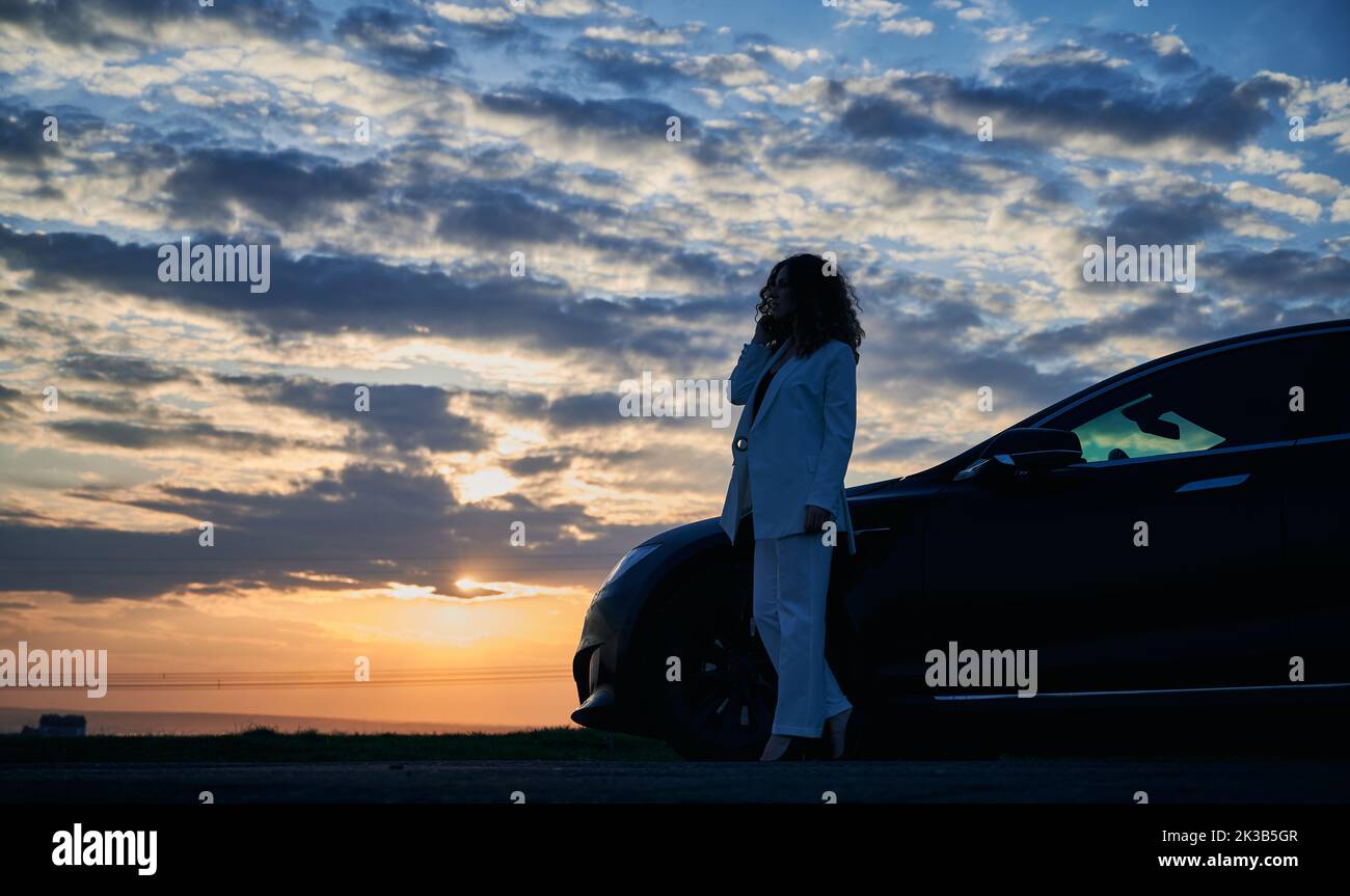 Beautiful sunset. Silhouette of girl standing on road, enjoying incredible sunset over horizon at cloudy sky. Luxury black car parked to her right. Stock Photo